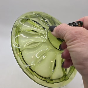 May include: A green glass lid with a decorative pattern of curved lines and oval shapes. The lid is held by a hand with dark nail polish. The glass has a slight yellow tint and is partially transparent.