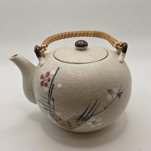 May include: A beige ceramic teapot with a woven handle and a lid. The teapot is decorated with floral designs in red, white, and black. The spout and handle attachments are brown.