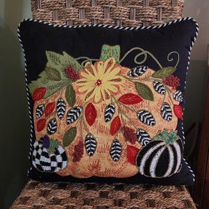 Op de afbeelding: Een decoratief vierkant kussen met een zwarte achtergrond en een gedetailleerd geborduurd pompoenontwerp. De pompoen is oranje met een gele bloem, groene bladeren en kralenaccenten. Twee kleinere pompoenen bevinden zich op de hoeken. Het kussen heeft een wit en zwart gestreepte rand.