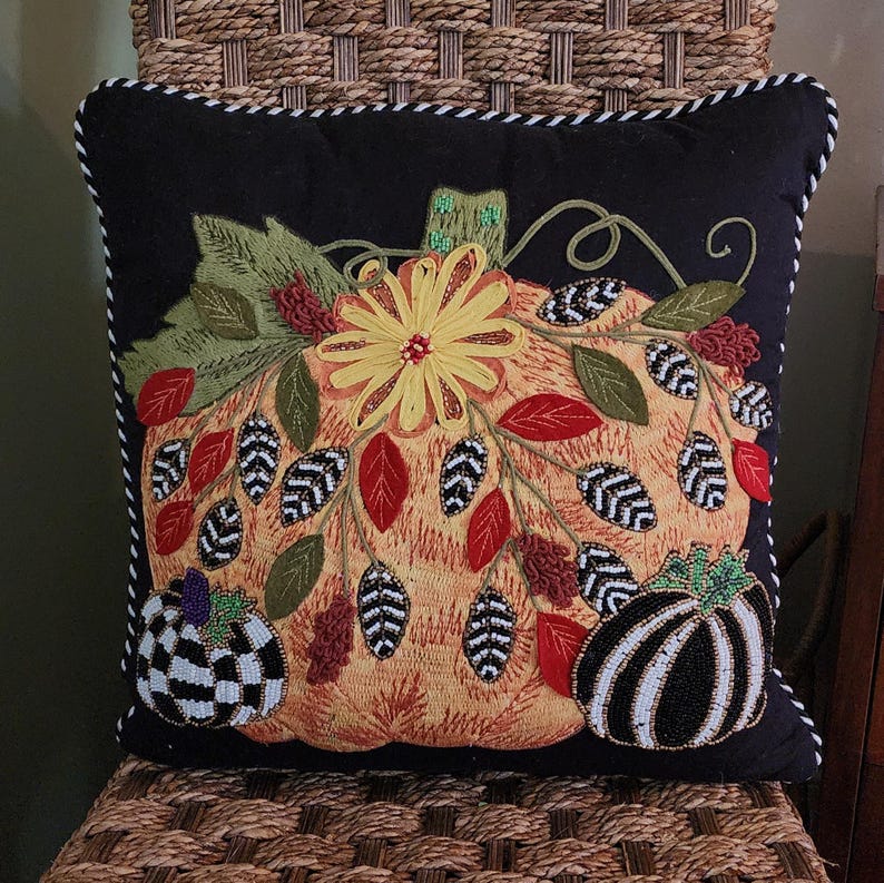 Op de afbeelding: Een decoratief vierkant kussen met een zwarte achtergrond en een gedetailleerd geborduurd pompoenontwerp. De pompoen is oranje met een gele bloem, groene bladeren en kralenaccenten. Twee kleinere pompoenen bevinden zich op de hoeken. Het kussen heeft een wit en zwart gestreepte rand.