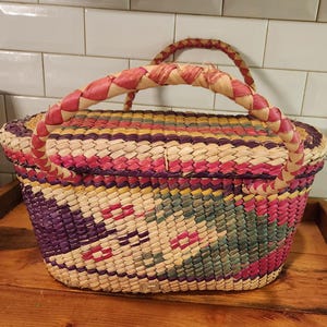 May include: A colourful woven basket with a lid and handle. The basket has a pattern of purple, pink, yellow, and green. The handle is wrapped in pink and cream-coloured material. The basket is on a wooden surface.