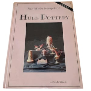 May include: A hardcover book titled The Collectors Encyclopedia of Hull Pottery by Brenda Roberts. The pale pink cover features a black-bordered central photograph displaying various ceramic vases and pitchers, with a black corner banner.