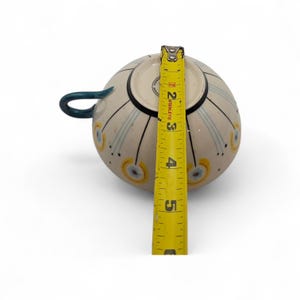 May include: A cream-colored ceramic teacup with a blue handle and decorative blue and yellow accents. The cup is approximately 3 inches tall, as measured by a yellow tape measure.