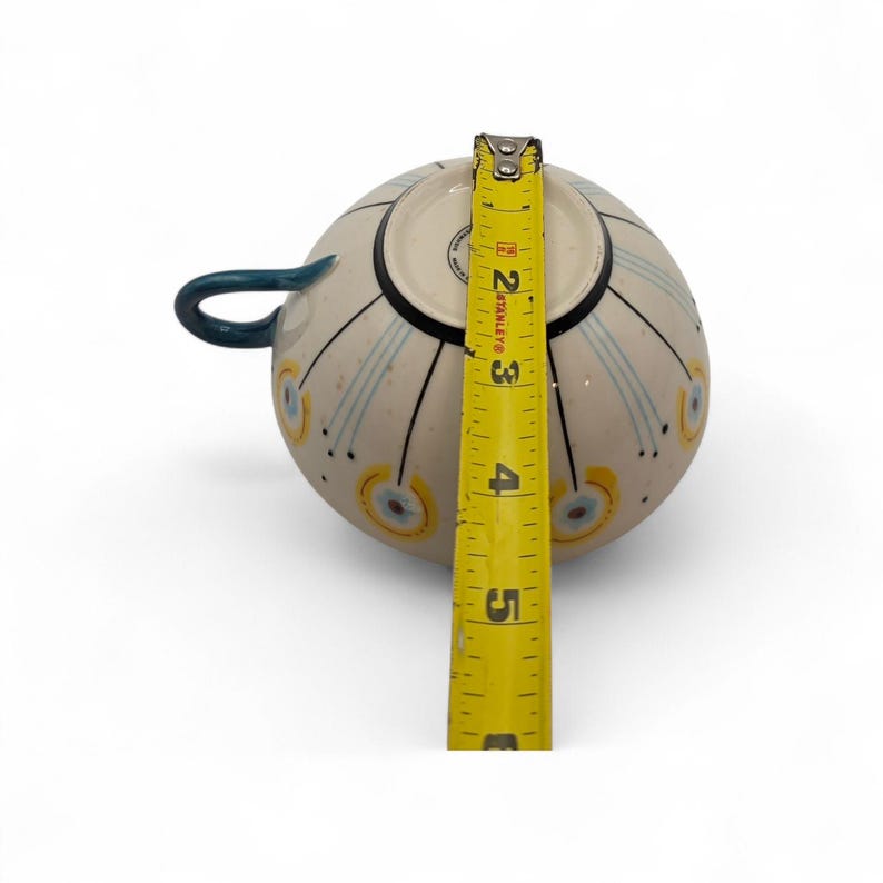 May include: A cream-colored ceramic teacup with a blue handle and decorative blue and yellow accents. The cup is approximately 3 inches tall, as measured by a yellow tape measure.