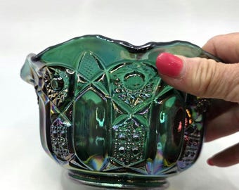 May include: A green carnival glass bowl with an iridescent finish, featuring a pressed pattern of geometric shapes, diamonds, and raised dots. The bowl has a scalloped rim and a reflective surface that displays hints of purple and gold tones.