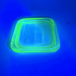 May include: A square, neon green glass dish with rounded corners. The dish is set against a vibrant blue background, creating a striking contrast. The glass appears translucent, with a subtle glow.