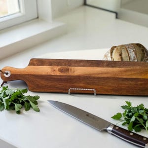 May include: A wooden cutting board with a handle, featuring a light and dark wood grain design. A stainless steel knife with a dark brown handle rests on the counter, along with fresh green herbs and a loaf of bread.