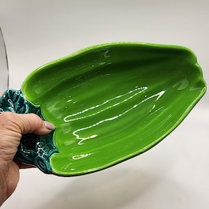 May include: A glossy, green ceramic serving dish in the shape of a leaf. The dish has a deep, curved interior and a textured, dark green handle. The bright green color and unique shape make it a decorative piece.