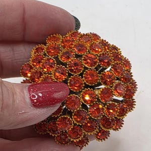 May include: A round, vintage brooch with a cluster of orange-red rhinestones. The stones are set in a gold-toned metal frame, creating a sparkling, eye-catching design. The brooch is a classic accessory, perfect for adding a touch of glamour to any outfit.