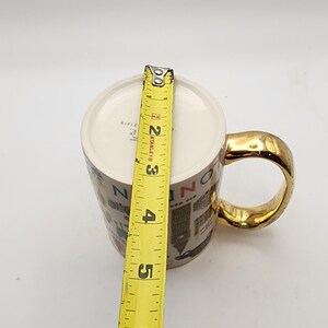 May include: A ceramic mug with a gold handle and rim, featuring a cityscape design. The mug is approximately 4 inches tall, as measured by a yellow tape measure. The mug's interior is white.