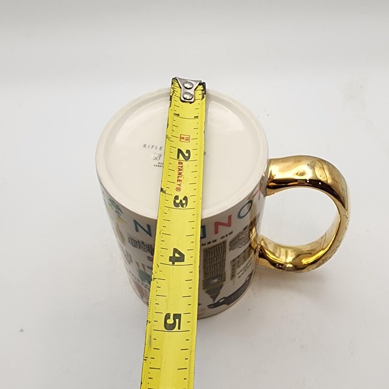 May include: A ceramic mug with a gold handle and rim, featuring a cityscape design. The mug is approximately 4 inches tall, as measured by a yellow tape measure. The mug's interior is white.