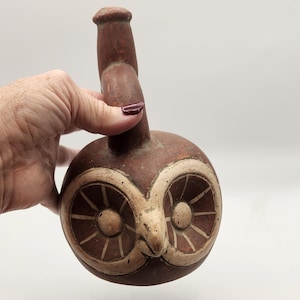 May include: A brown ceramic owl-shaped vessel with a long neck and a round body. The owl's face is detailed with large, white-rimmed eyes and a prominent beak. The vessel is held by a hand against a white background.