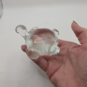May include: A clear glass turtle figurine with a rounded body, head, and four flipper-like appendages. The turtle is held in a hand, showcasing its transparent design. The figurine is a decorative item.