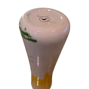 May include: A white vase with green and yellow accents. The vase has a rounded top and a long, slender body. The base of the vase is yellow, and the top is white with green leaf-like designs. The vase is likely made of ceramic or porcelain.