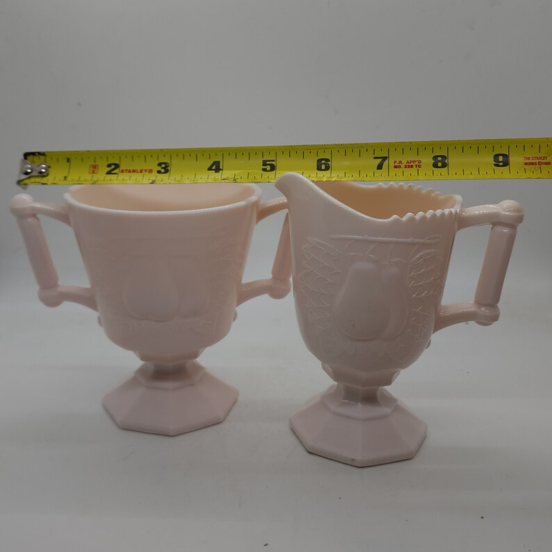 K&ouml;nnte beinhalten: Ein rosa Zuckerdosen- und Sahne-Set aus Glas. Die Zuckerdose hat zwei Griffe und einen Sockel. Der Sahneaufgie&szlig;er hat einen Ausguss und einen Griff. Beide Teile haben ein Birnen-Design. Die Zuckerdose ist ca. 7.6 cm breit.