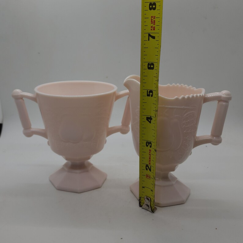 K&ouml;nnte beinhalten: Ein hellrosa Vintage-Zucker- und Sahne-Set. Die Zuckerdose hat zwei Griffe und einen Sockel. Der Sahnegie&szlig;er hat einen Ausguss und einen Griff. Der Sahnegie&szlig;er ist etwa 13 cm hoch. Beide Teile haben ein florales Design.