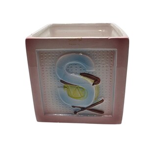 May include: A square ceramic container featuring a pink border and a textured white center panel. A large, light blue embossed letter S is centered on the front, accented by a small yellow drum graphic and two crossed brown drumsticks below it.