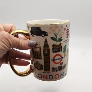 May include: Cream-colored ceramic mug with a gold handle and rim, featuring London landmarks like Big Ben, a black taxi, and the London Underground logo. The word "LONDON" is printed at the bottom of the mug.