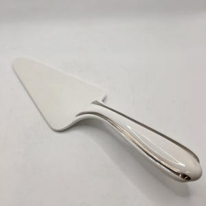 May include: A white ceramic cake server with a triangular blade and a smooth, rounded handle. The handle features a thin, metallic silver trim that runs along the edges, providing a subtle contrast to the glossy white finish of the utensil.