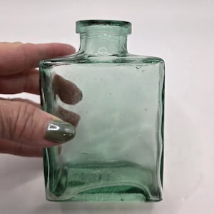 May include: A square, antique-style glass bottle with a light green tint. The bottle has a rectangular body and a short, cylindrical neck. The glass appears slightly textured. The bottle is held by a hand.