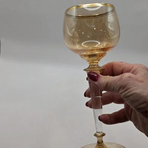 May include: A vintage amber-colored wine glass with a gold rim and a clear, textured stem. The glass has a rounded bowl and a decorative base. The stem has a textured, vertical pattern. The glass is held by a person.