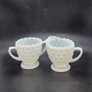 May include: A set of two white milk glass creamer and sugar bowls with a textured, bubbled design. The bowls have a scalloped edge and a footed base.