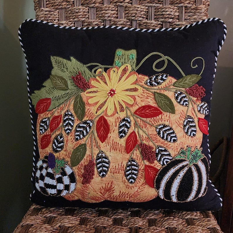 Op de afbeelding: Zwart vierkant decoratief kussen met een geborduurd pompoenontwerp. De pompoen is oranje met een gele bloem en groene bladeren. Twee kleinere pompoenen staan ​​onderaan, een geblokt en een gestreept.