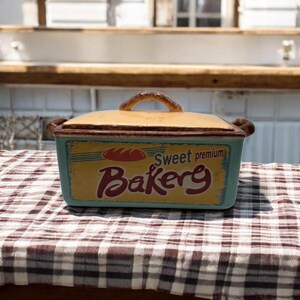 May include: A rectangular ceramic bread box with a lid. The box is light blue with a yellow label that reads "Sweet premium Bakery" in red and brown lettering. The lid is tan with a brown handle.