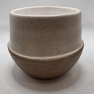May include: A ceramic planter with a two-tone finish. The upper section features a glossy, off-white crackle glaze with dark speckles, while the lower section has a textured, matte, sandy-brown finish separated by a distinct horizontal ridge.
