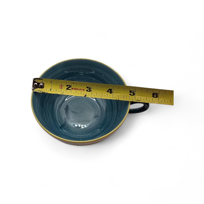 May include: A teal ceramic teacup with a black handle and a gold rim is measured with a yellow tape measure. The tape measure shows the cup is approximately 6 inches in diameter. The cup has a glossy finish.