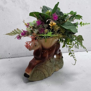 May include: A decorative ceramic vase in the shape of a deer, holding an arrangement of artificial flowers and greenery. The vase is brown, beige, and green, with pink, purple, and yellow flowers. The deer is in a leaping pose.