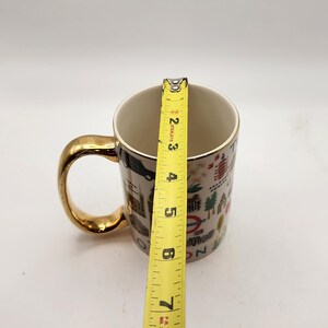 May include: A white ceramic mug with a gold handle and rim. The mug is decorated with London landmarks and icons. The mug is approximately 4 inches tall, as measured by a yellow tape measure.