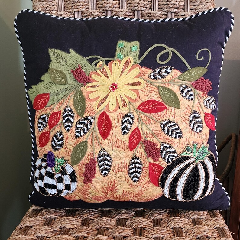 Op de afbeelding: Decoratief vierkant kussen met een zwarte achtergrond en een groot geborduurd pompoenontwerp. De pompoen is oranje met een gele bloem, groene bladeren en rode en zwarte accenten. Het kussen heeft een wit en zwart gestreepte rand.