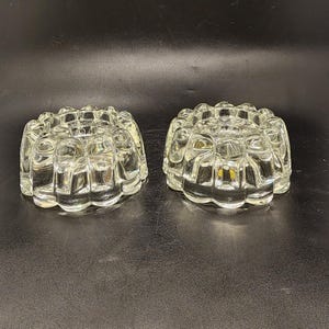 May include: Two clear glass candle holders with a decorative, textured design. The holders are round and have a scalloped edge.