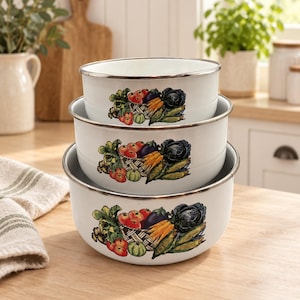 May include: A set of three white enamel nesting bowls stacked vertically. Each bowl features a silver-toned rim and a colorful graphic print of a woven basket filled with fresh garden vegetables, including tomatoes, carrots, corn, and eggplant.
