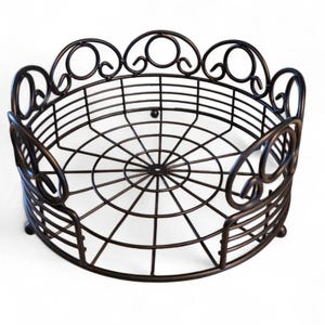 May include: A round, bronze-colored metal basket with an openwork design. The basket features a decorative scalloped edge and a radial pattern on the base. The sides have curved handles. This decorative basket is suitable for various home decor uses.