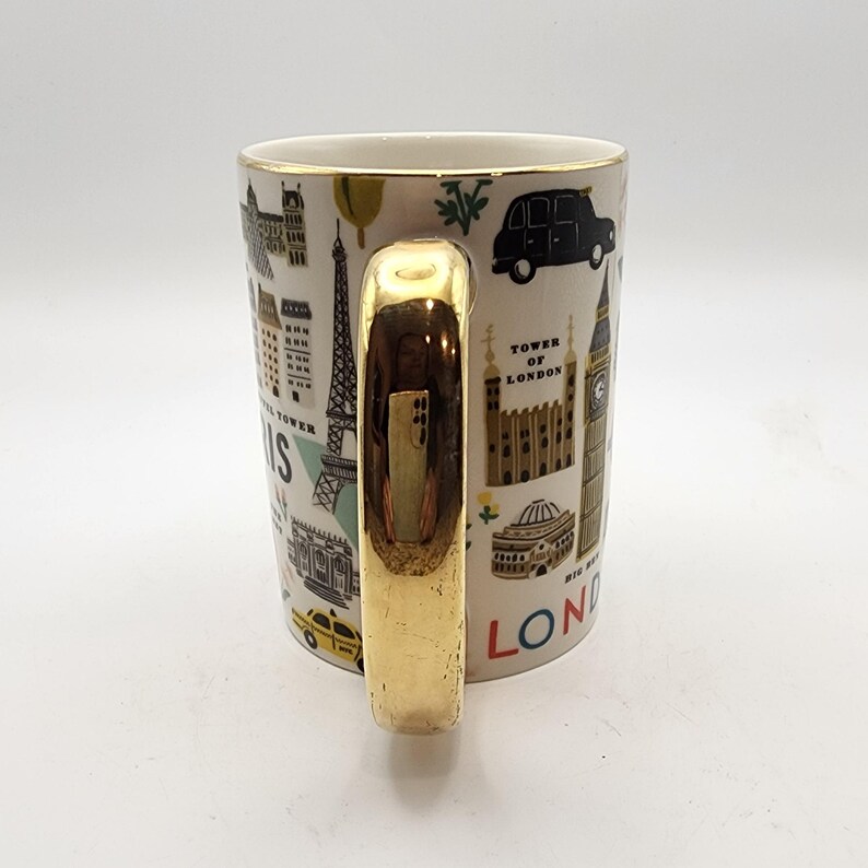 May include: White ceramic mug with a gold handle and rim, featuring illustrations of famous landmarks. The mug includes images of the Eiffel Tower, Big Ben, and a black car. Text on the mug reads "PARIS" and "LONDON".