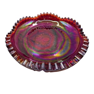 May include: A round, iridescent carnival glass bowl featuring a deep red hue with purple and gold highlights. The bowl has a scalloped, serrated edge and a central pressed geometric star pattern, showcasing a reflective, metallic-like finish.