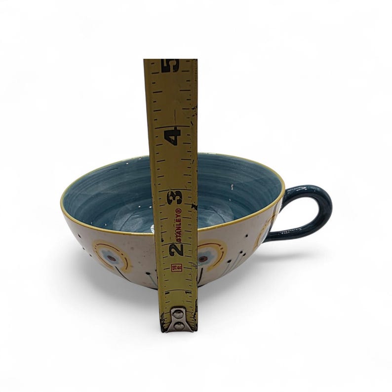 May include: A ceramic teacup with a blue interior and a cream-colored exterior. The cup has a black handle and is approximately 3 inches (7.6 cm) tall. The exterior features a floral design with yellow and black accents.