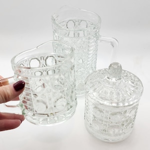 May include: A clear glass pitcher, mug, and lidded jar set. The glass pieces have a textured, geometric pattern. The pitcher has a handle and a spout. The mug has a handle. The jar has a round shape and a knob on the lid. The set is on a white background.