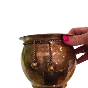 May include: A round, hammered copper pot featuring a wide, flared rim and a decorative rope-like metal trim encircling the upper section. Two twisted metal cords hang from the rope trim, each ending in a small, smooth, spherical metal bead.