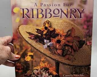 May include: A book cover titled "A Passion For Ribbonry" featuring a straw hat adorned with colorful ribbon flowers. The cover has a floral background and the author's name, Camela Nitschke, is visible.