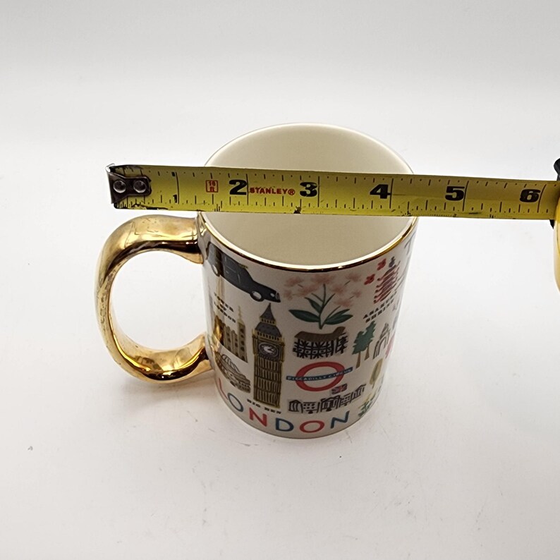 May include: A white ceramic mug with a gold handle and rim. The mug features a London-themed design with iconic landmarks and the word "LONDON". The mug's opening measures approximately 3.5 inches in diameter.