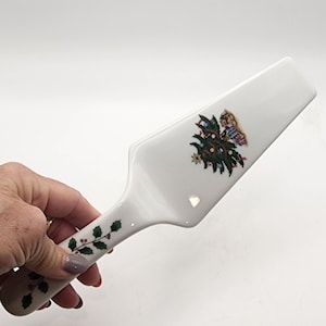 May include: White ceramic pie server with a Christmas tree and holly berry design. The server features a wide, flat blade and a handle decorated with green holly leaves and red berries. A festive holiday kitchen accessory.