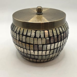 May include: A decorative jar with a lid. The jar is covered in small, rectangular, mirrored tiles in shades of silver and gray. The lid and small knob are a bronze color. The jar is round and has a classic shape.