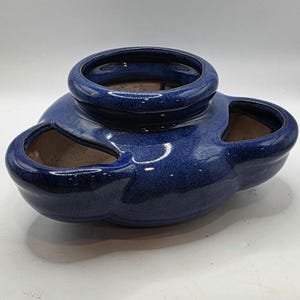 May include: A glazed, cobalt blue ceramic strawberry pot. The pot has a round top opening and three side openings. The exterior has a speckled, glossy finish. The interior is unglazed and a light brown color. The pot is designed for planting strawberries.