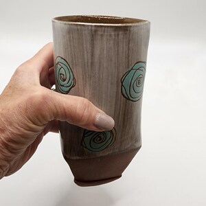 May include: A handmade ceramic tumbler with a brown base and rim, and a light gray body. The body features three turquoise spiral designs. The tumbler is held by a person.