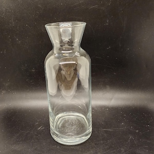 May include: A clear glass pitcher with a wide mouth and a narrow neck. The pitcher is empty and has a smooth, polished surface.