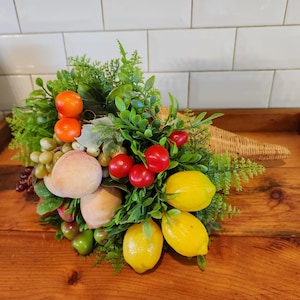 May include: A woven cornucopia overflowing with faux fruit and greenery. The arrangement includes bright yellow lemons, red cherries, orange mandarin oranges, green and purple grapes, and peaches. The cornucopia is set on a wooden surface.