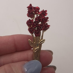 May include: A gold-toned metal brooch featuring a bouquet of red roses. The roses are clustered together, with detailed petals and a glossy finish. The brooch has a classic design, suitable for adding a touch of elegance to any outfit.